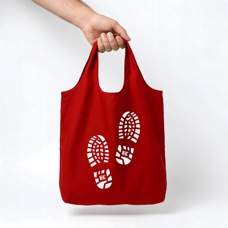A hand holding a red Kramp tote bag, with two white Kramp boot print illustrations on the front. 