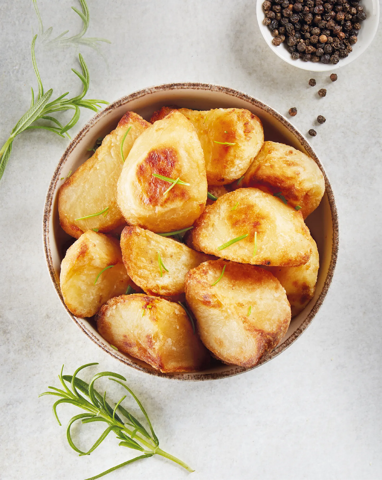 Overhead photography of roasted potatoes in a rustic bowl, surrounded by sprigs of rosemary and a bowl of black pepper.