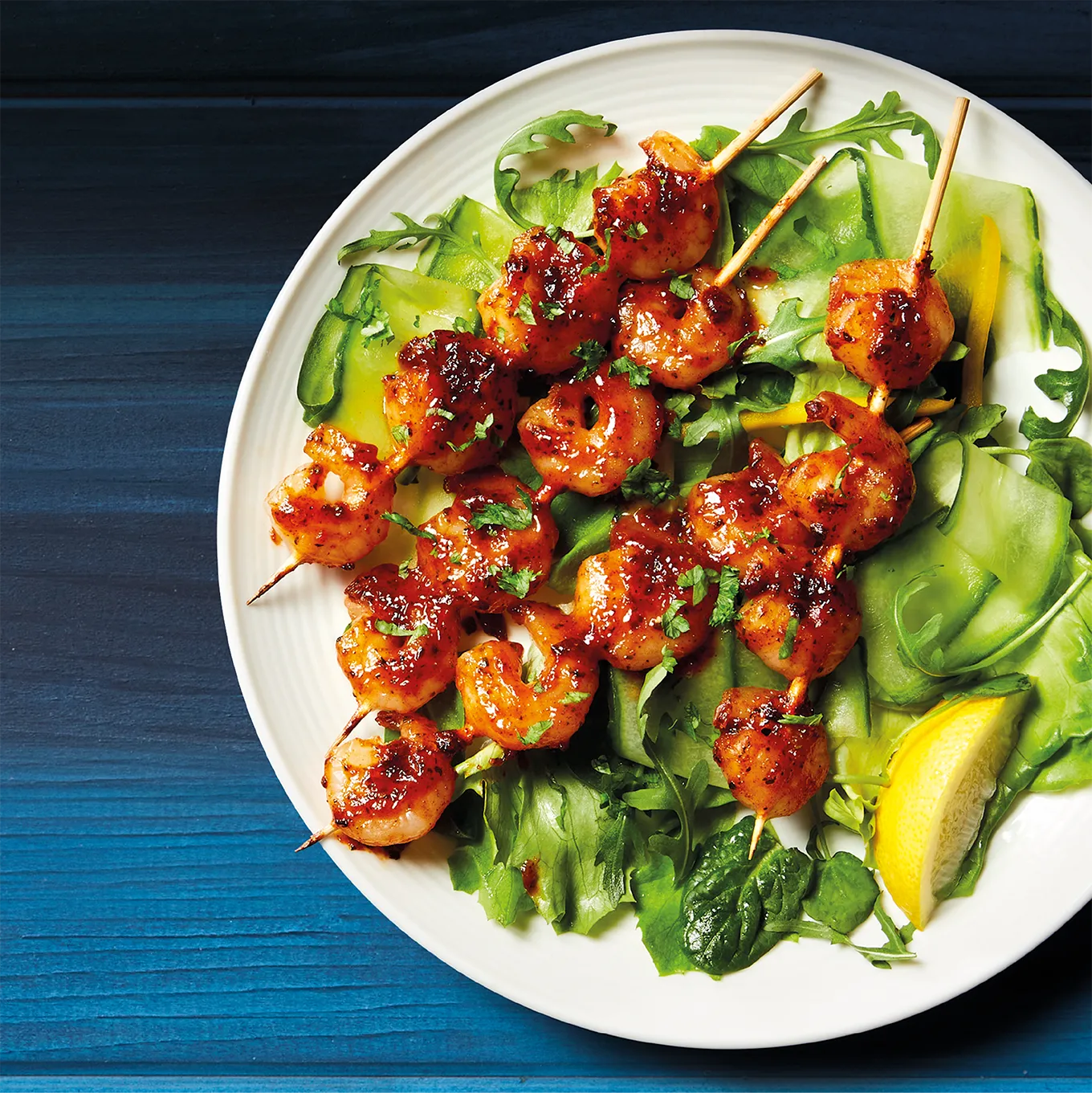 Styled food photography of teriyaki prawn skewers served with salad and a lemon wedge. Shot on a textured white plate, against a rustic, blue wooden background.
Shot in-house by Stocks Taylor Benson.