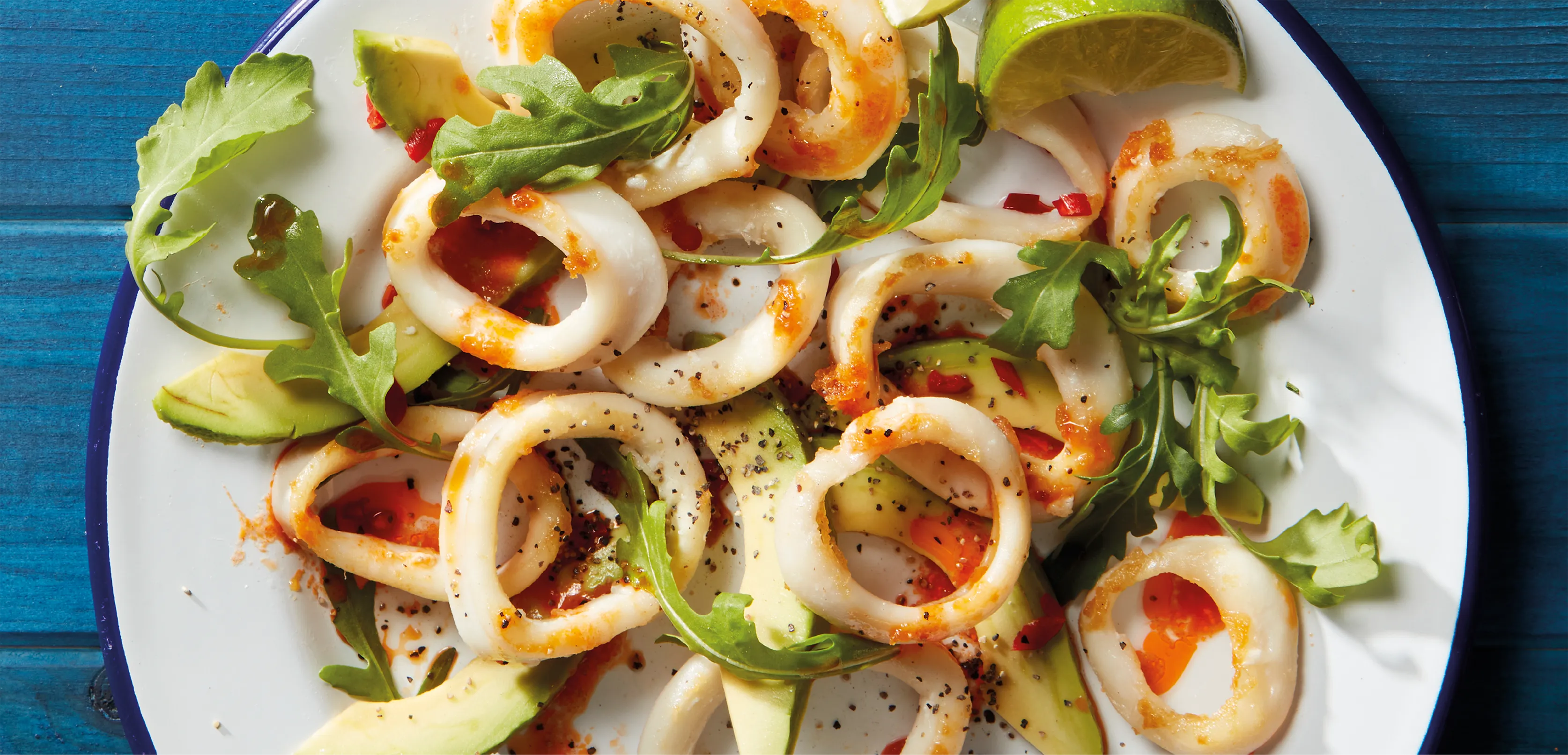 Styled food photography of squid rings, served with an avocado and rocket salad. Shot on a white plate, against a rustic, blue wooden background. 
Shot in-house by Stocks Taylor Benson.